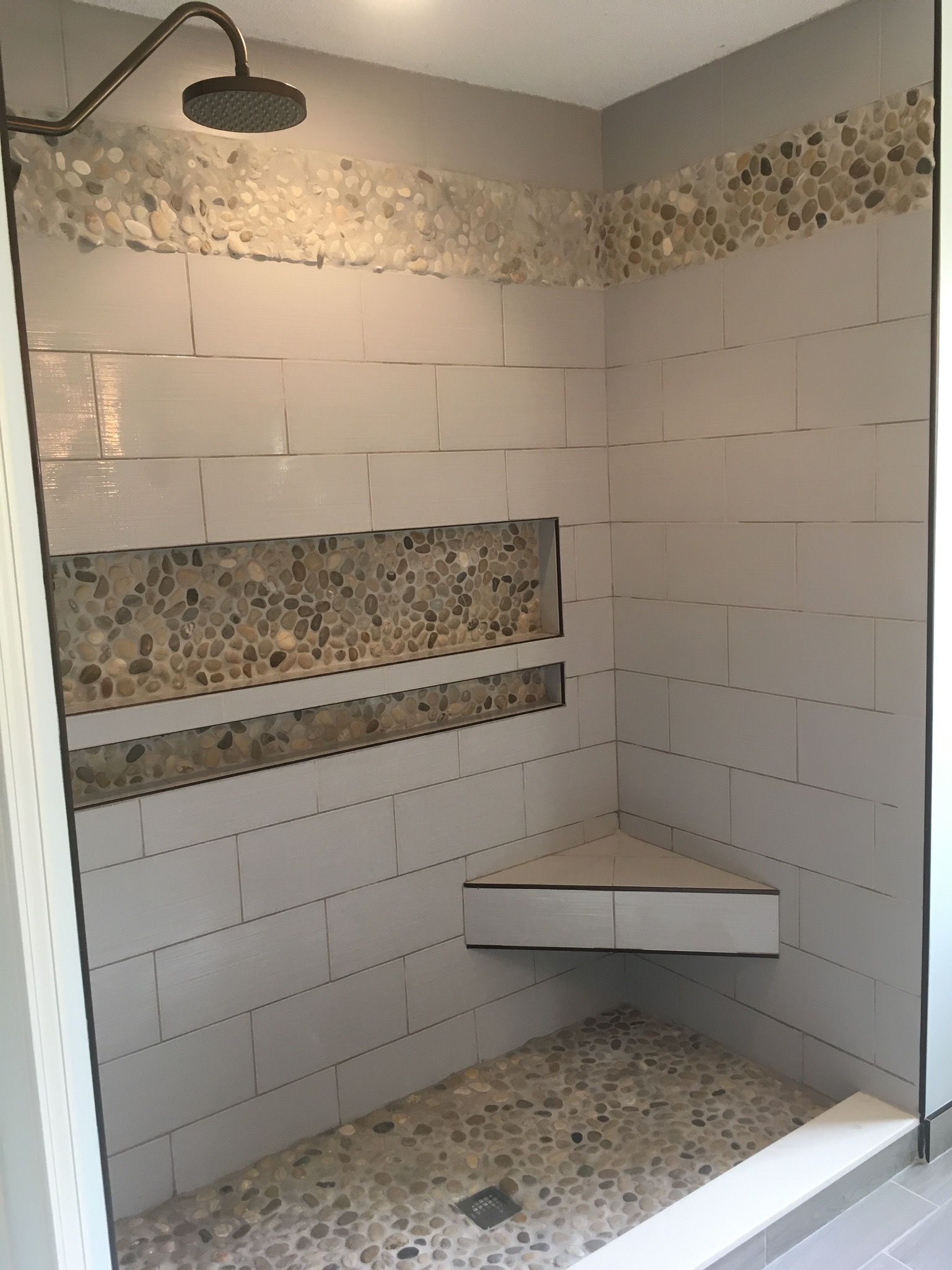 Shower with light gray tile walls, pebble floor, and corner shelf. Accents with light and dark pebbles.