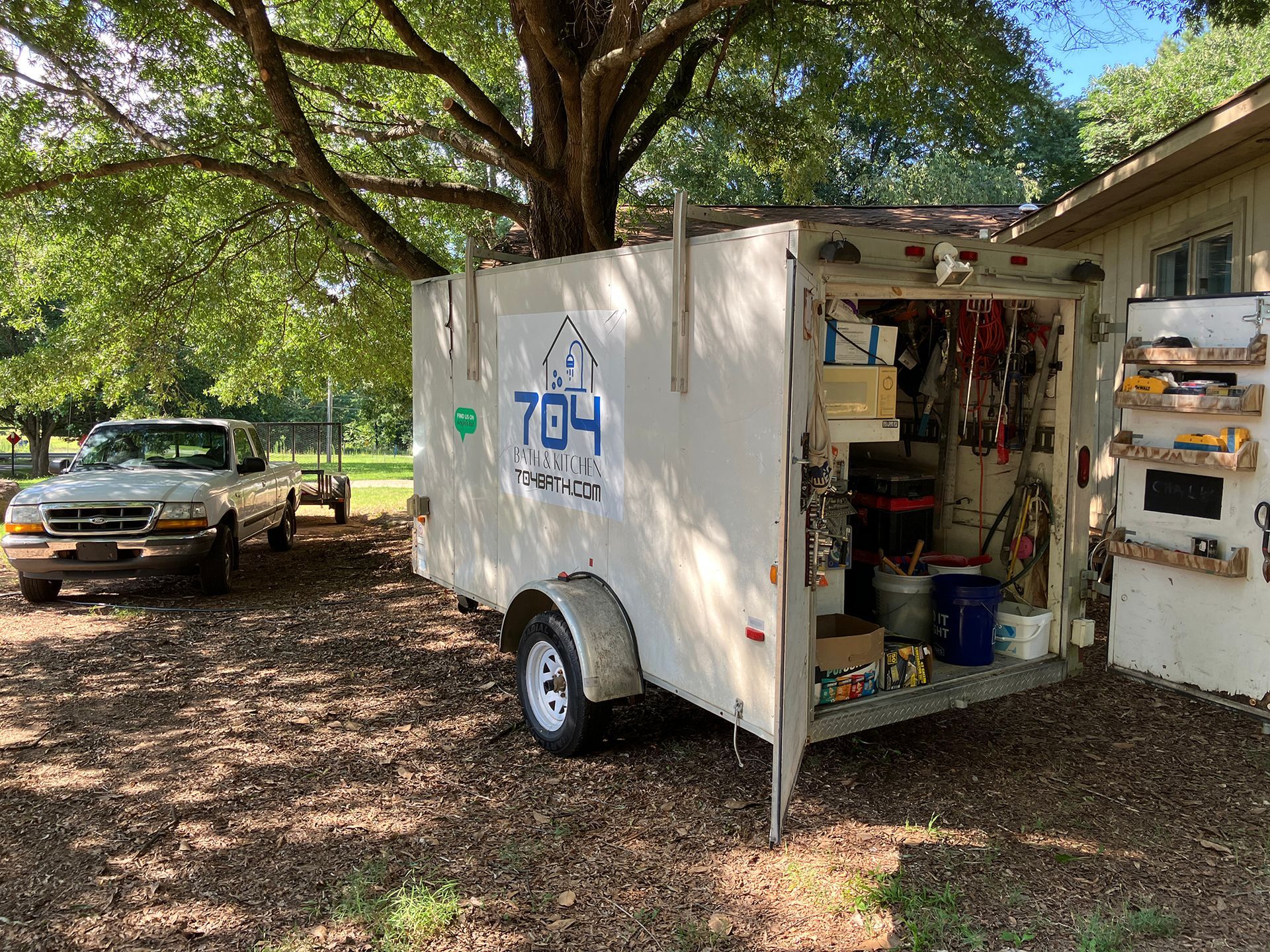 White trailer with tools open next to a building and a truck. Tree overhead.
