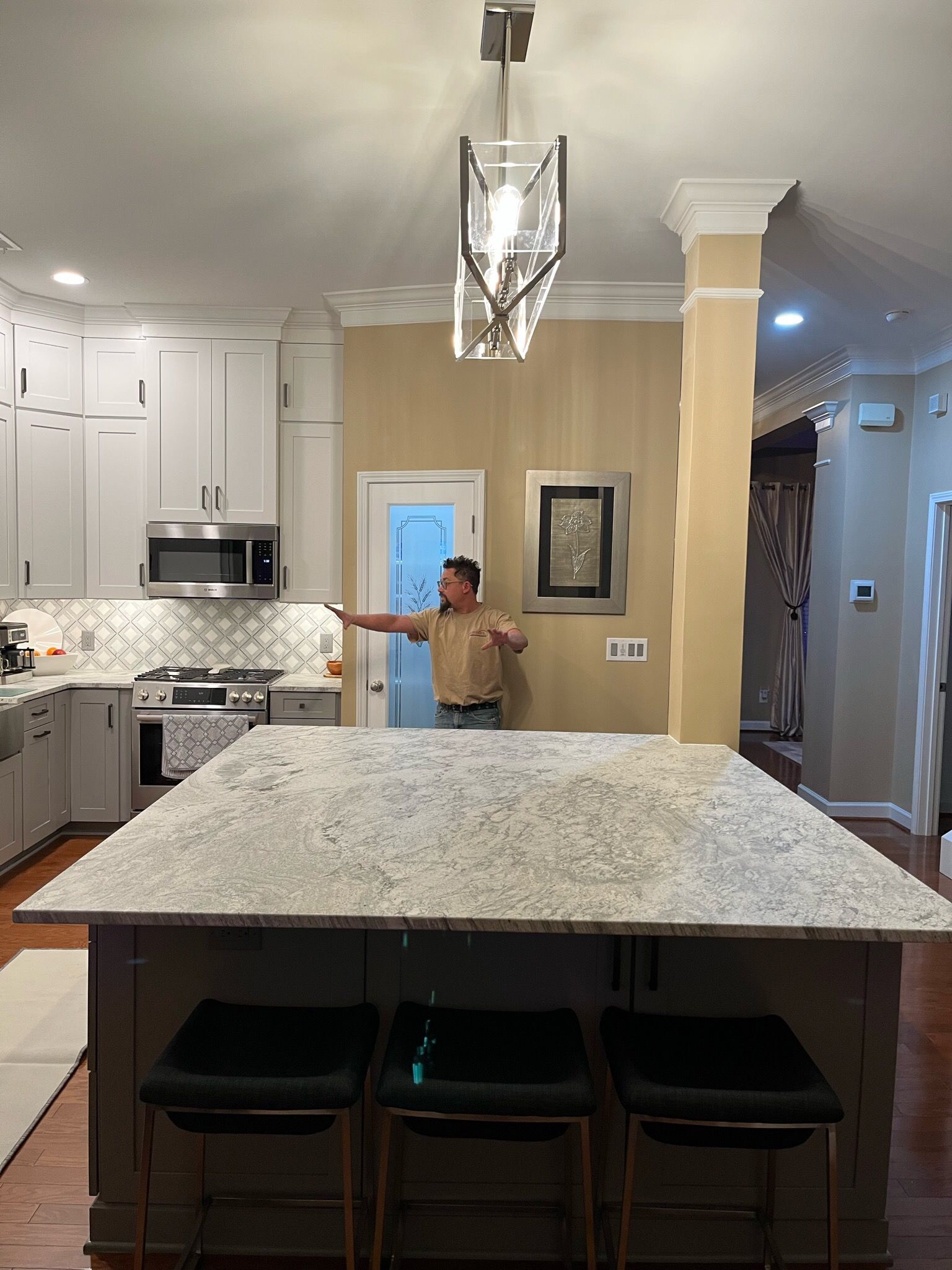 Man in kitchen with large island, extending arm toward a door. Light cabinets, light granite countertop, and three bar stools.