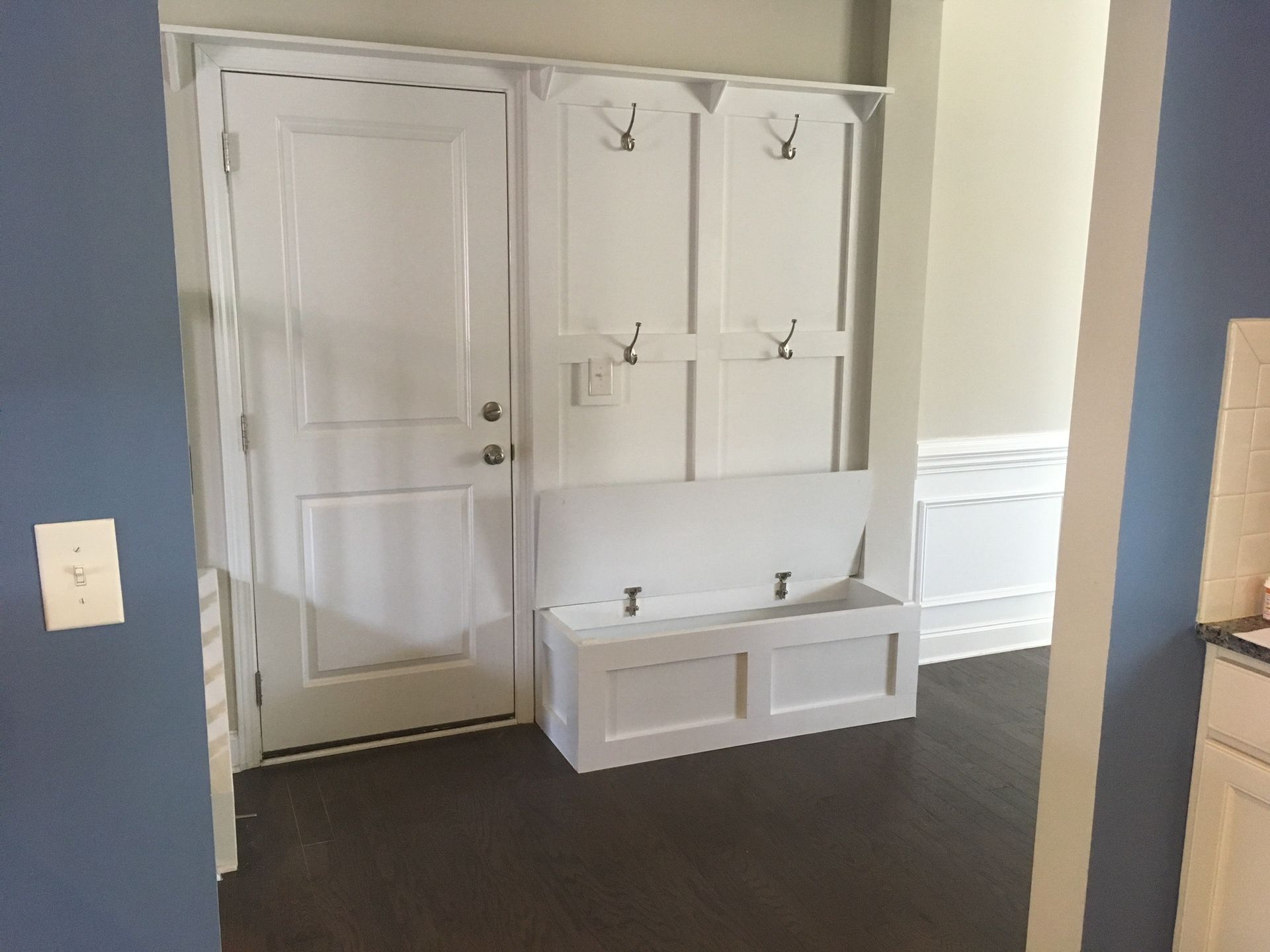 White entryway built-in with hooks, bench, and door, against dark wood floor and blue and beige walls.