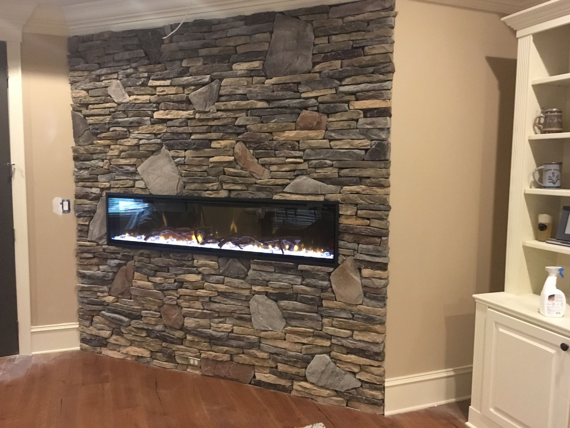 Stone wall with a linear electric fireplace, beside a built-in white shelf.