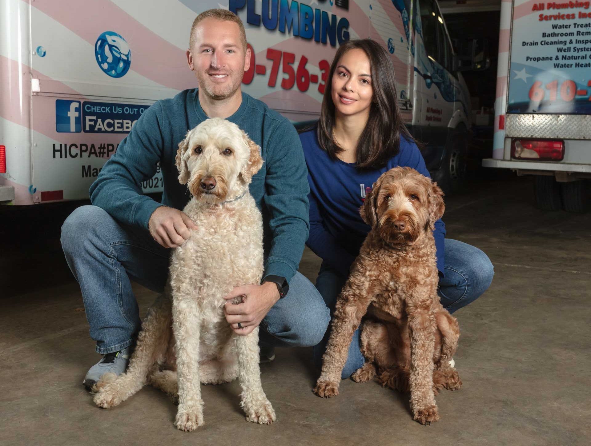 A man and a woman pose with two curly-haired dogs in front of a service vehicle with plumbing branding.