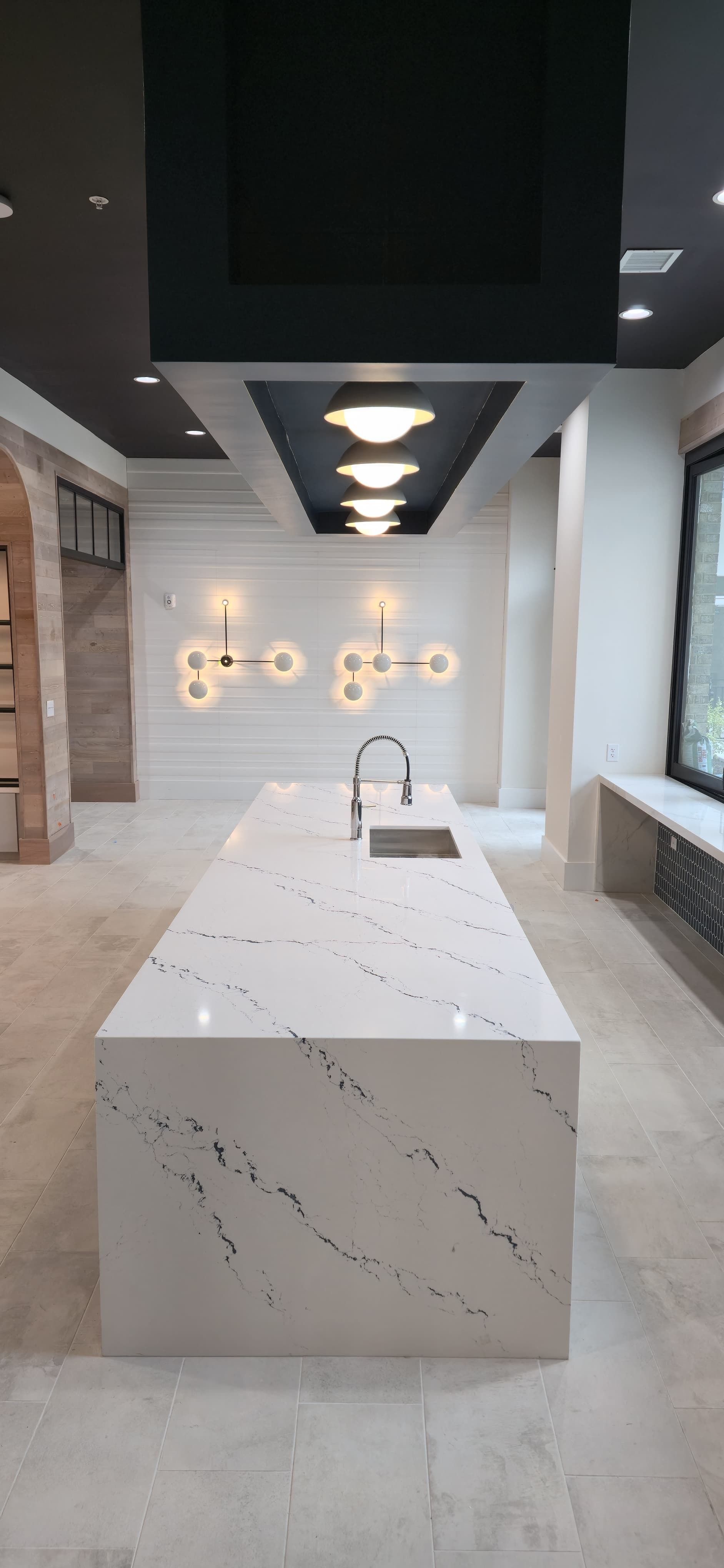 Modern white kitchen with marble island, black ceiling accents, pendant lights, and a sink by the window