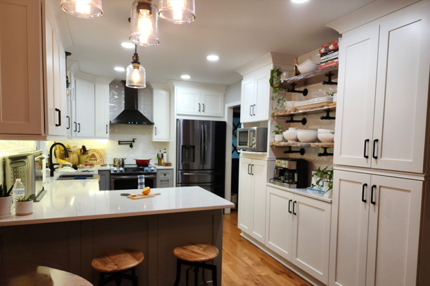 Modern white kitchen with wooden floors, an island with bar stools, open shelving, and a stainless steel refrigerator.
