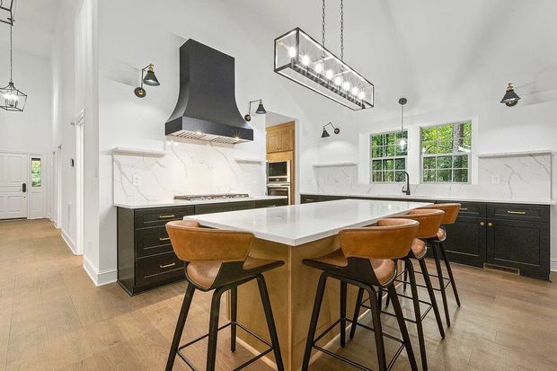 A modern kitchen with black cabinets, a large white island with brown stools, and high ceilings with pendant lighting.