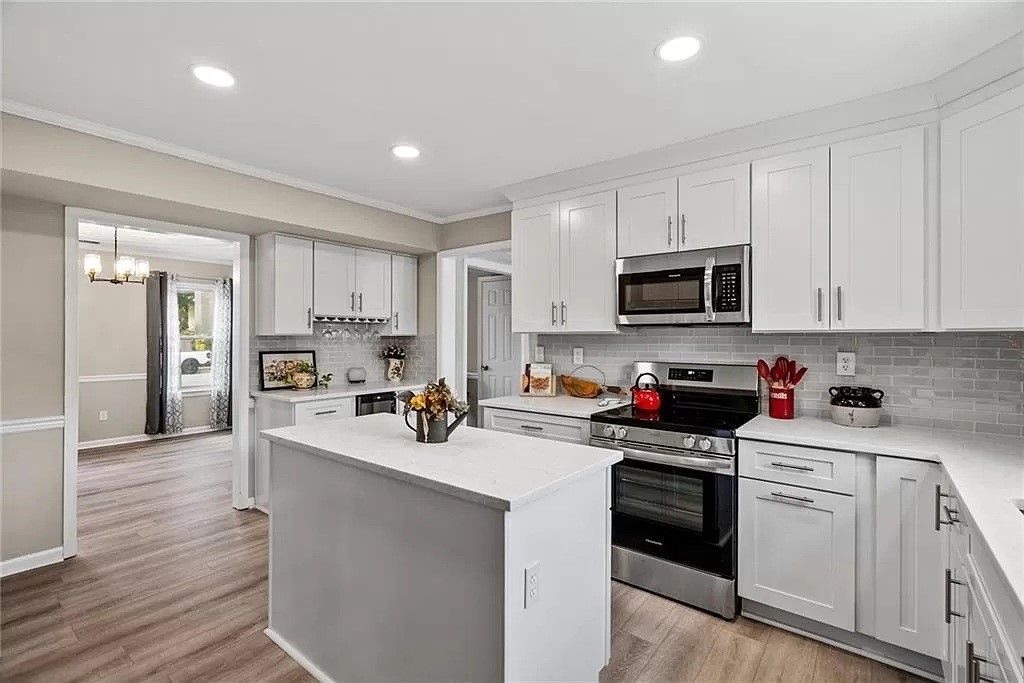 Modern white kitchen with island, stainless steel appliances, and adjacent room visible.