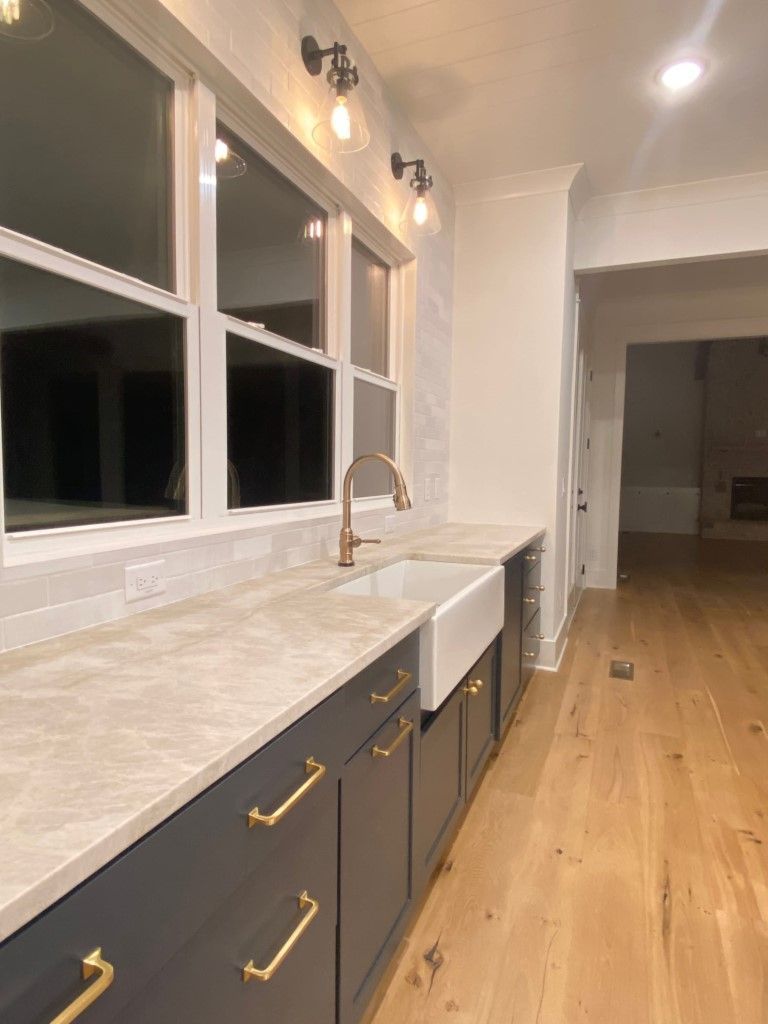 Kitchen with gray cabinets, gold hardware, white farmhouse sink, light countertops, and large windows.