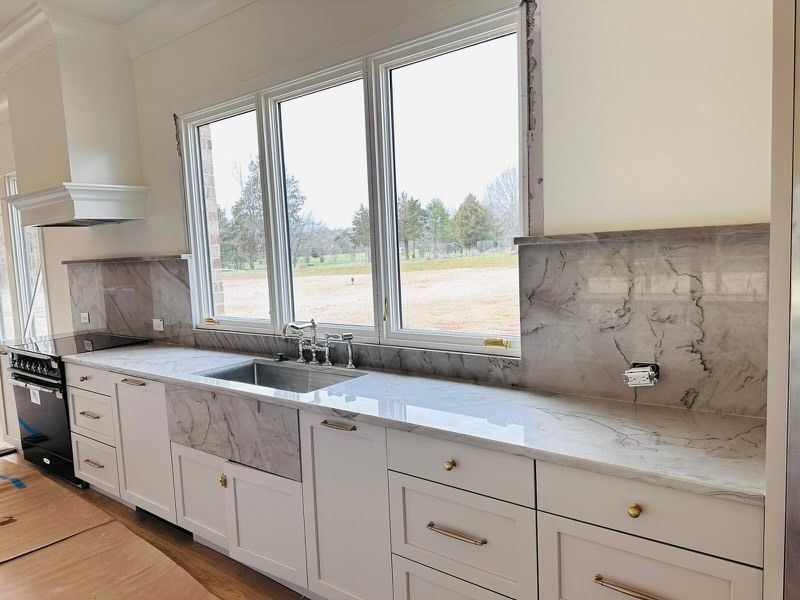 Kitchen with white cabinets, marble countertops, stainless steel sink, and large windows overlooking a green field.