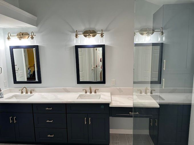 Bathroom with navy blue cabinets, white countertops, two sinks, mirrors, and gold light fixtures.