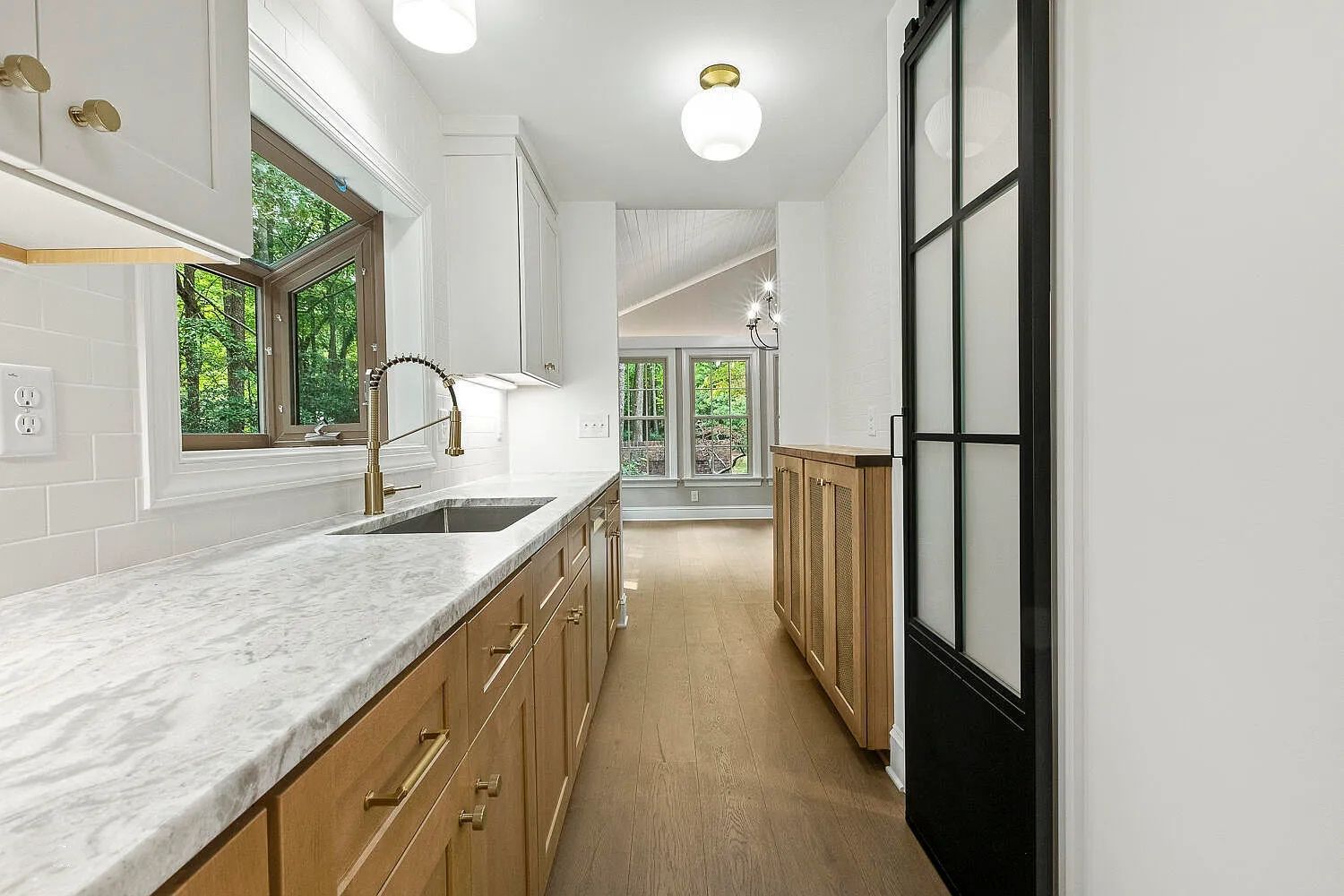 Long, narrow kitchen with wood cabinets, white countertops, and a window overlooking greenery.