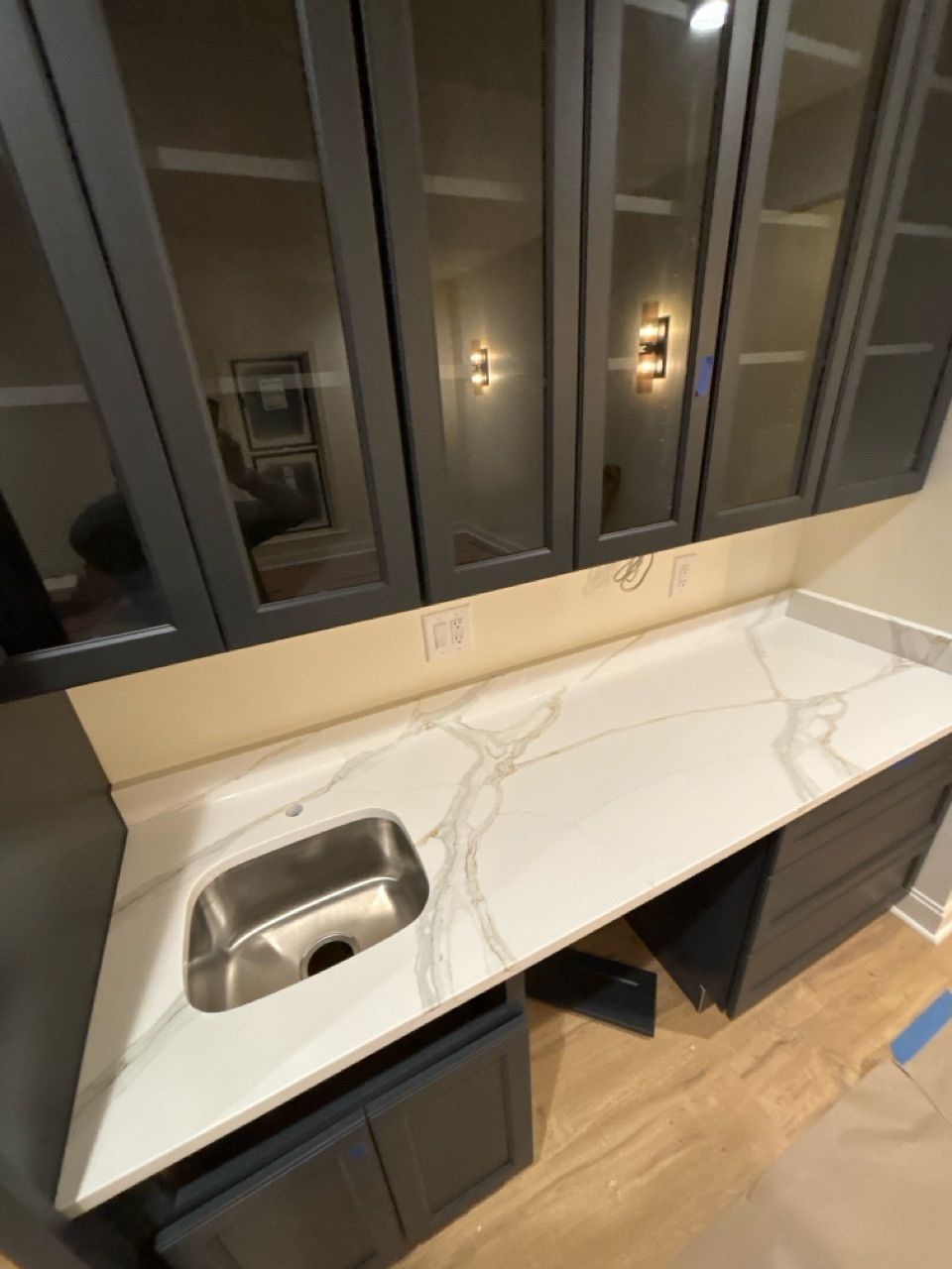 Modern bar area with white marble countertop, stainless steel sink, dark cabinets, and glass-front upper cabinets.