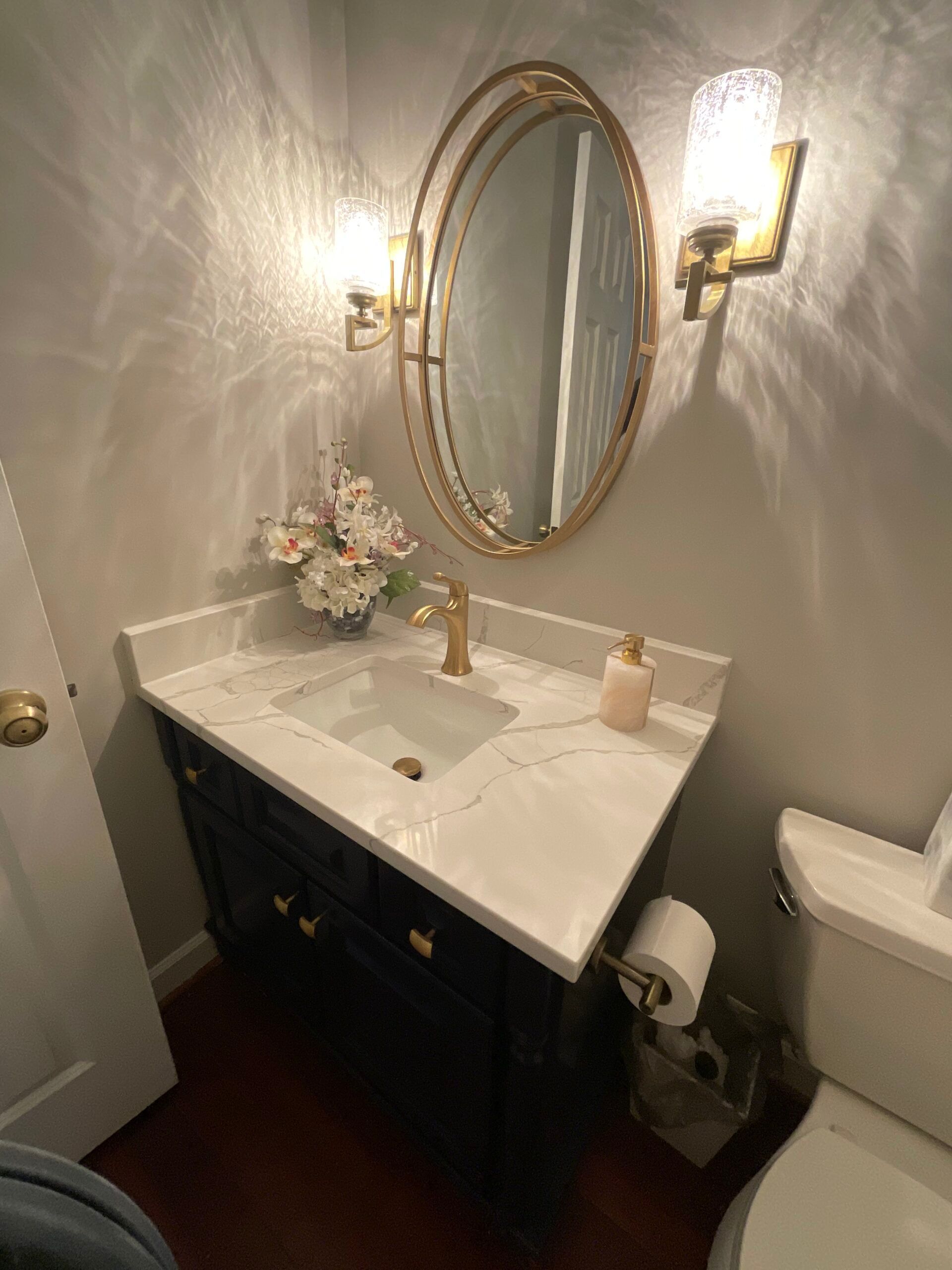 Powder room with gold-framed mirror, sconces, navy vanity, marble countertop, and toilet.