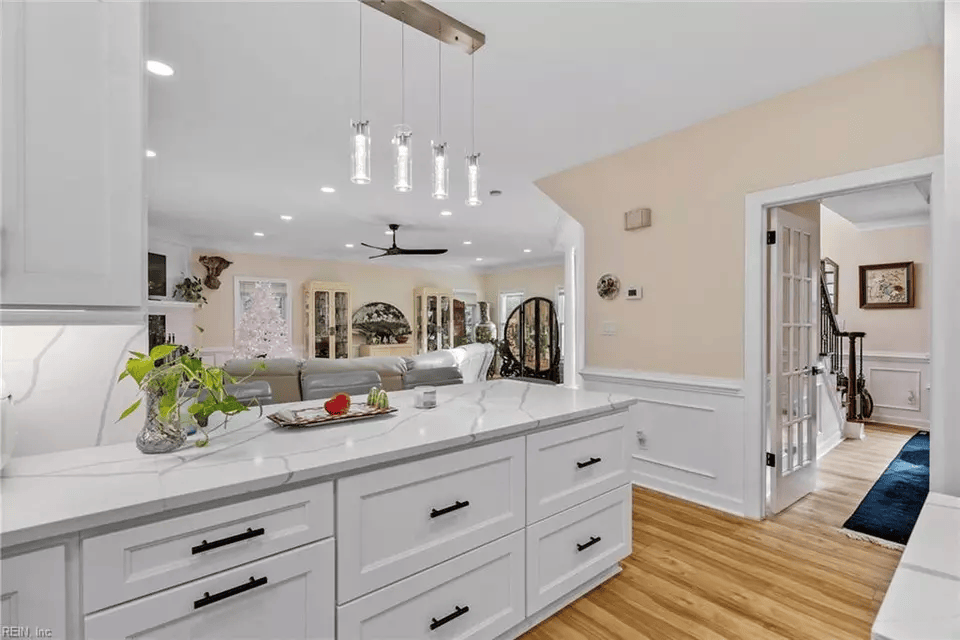 White kitchen with island, view into living area and hallway with staircase.
