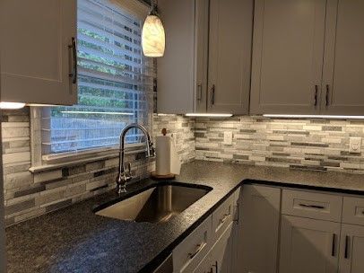 Kitchen corner with light gray cabinets, dark countertops, a stainless steel sink, and a decorative backsplash.