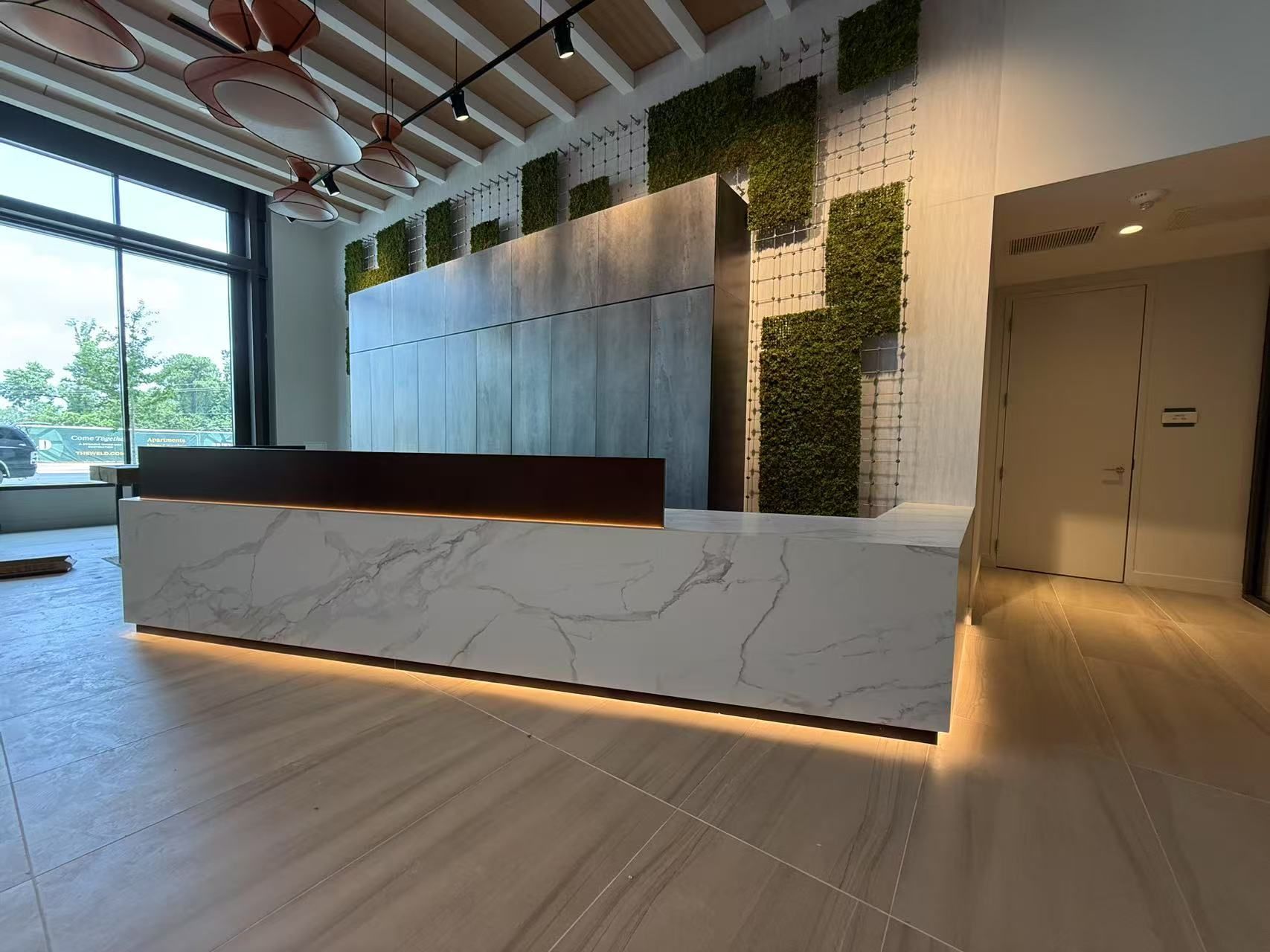 Reception desk with marble-look counter and wood accents. Green moss wall and large windows in background.