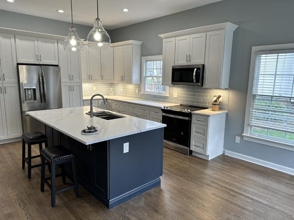 Modern white kitchen with dark blue island, stainless steel appliances, and wooden floors.
