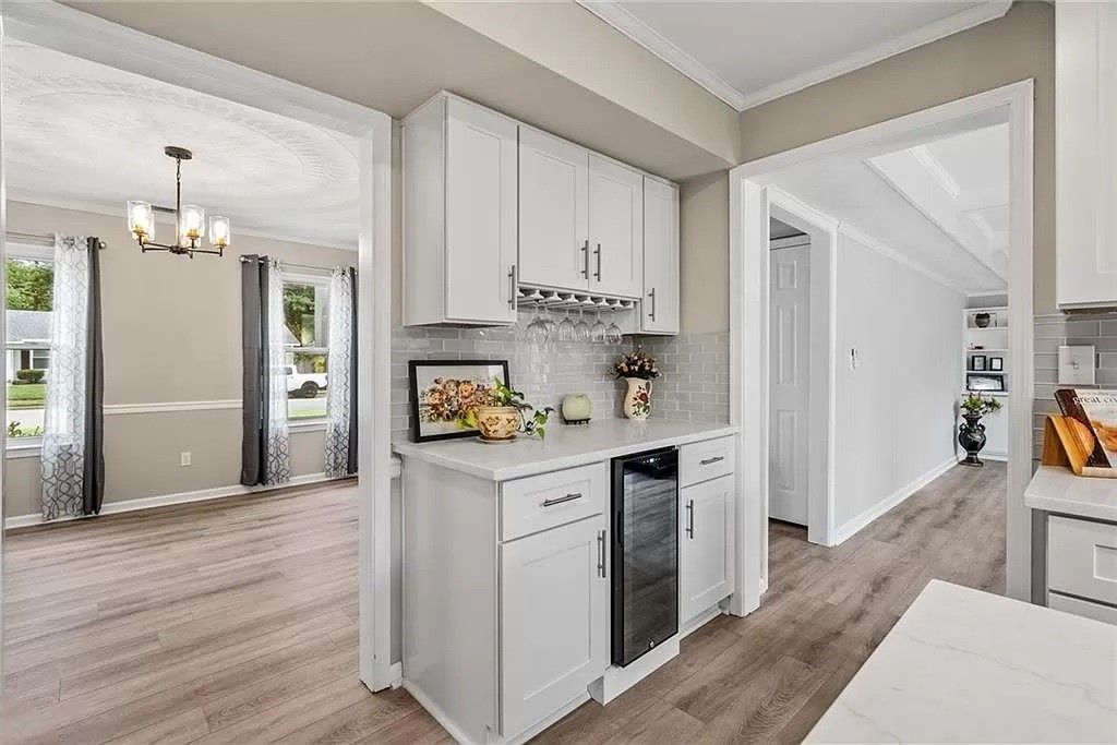 White cabinets with a wine cooler, framed by doorways, open to a living area with windows and a dining area.