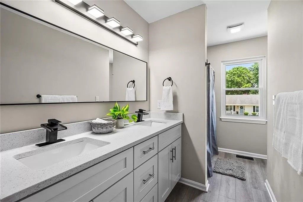 Bathroom with gray walls, light fixtures above a large mirror, dual sinks, and a window.