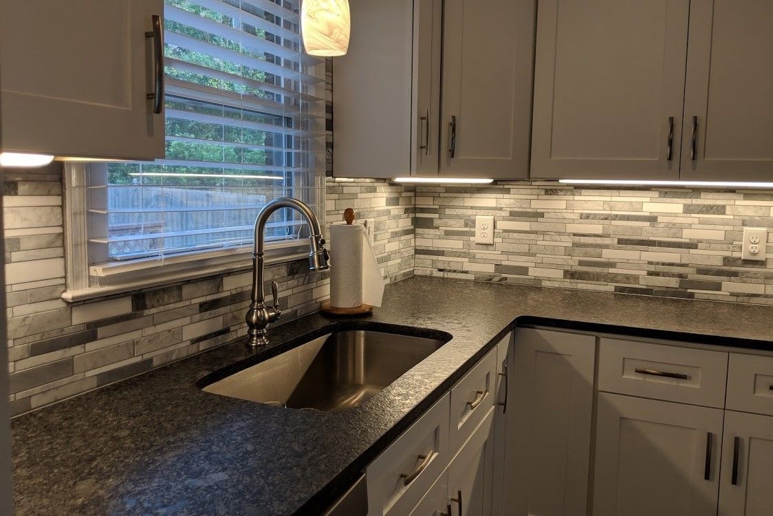Kitchen with gray brick backsplash, stainless steel sink, and white cabinets under bright lights.