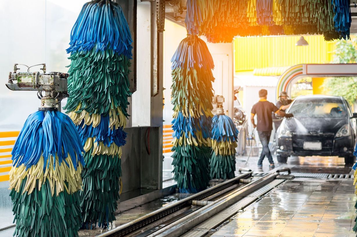 Car in a car wash, surrounded by blue, green, and yellow brushes. Two people, one spraying the car with water.