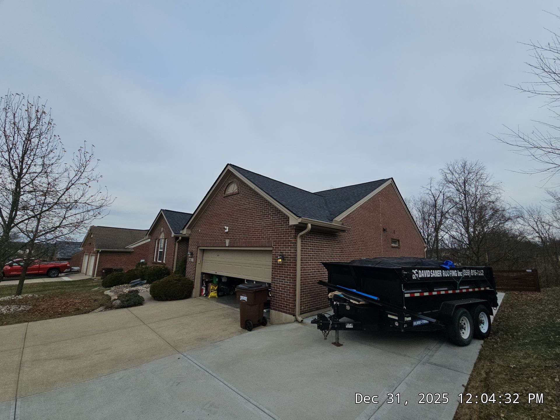 Brick house with open garage door; black dumpster trailer in driveway. Overcast day.