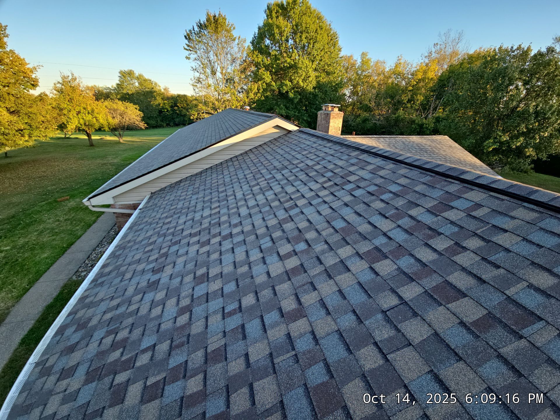 Roof with dark gray and blue shingles, chimney, and surrounding trees.