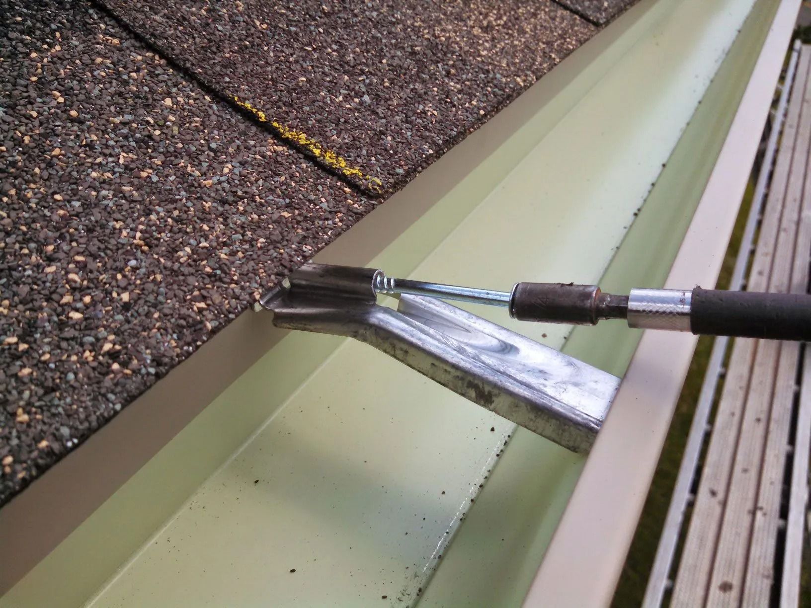 A gutter being cleaned with a special tool. Roof shingles and a section of scaffolding visible.