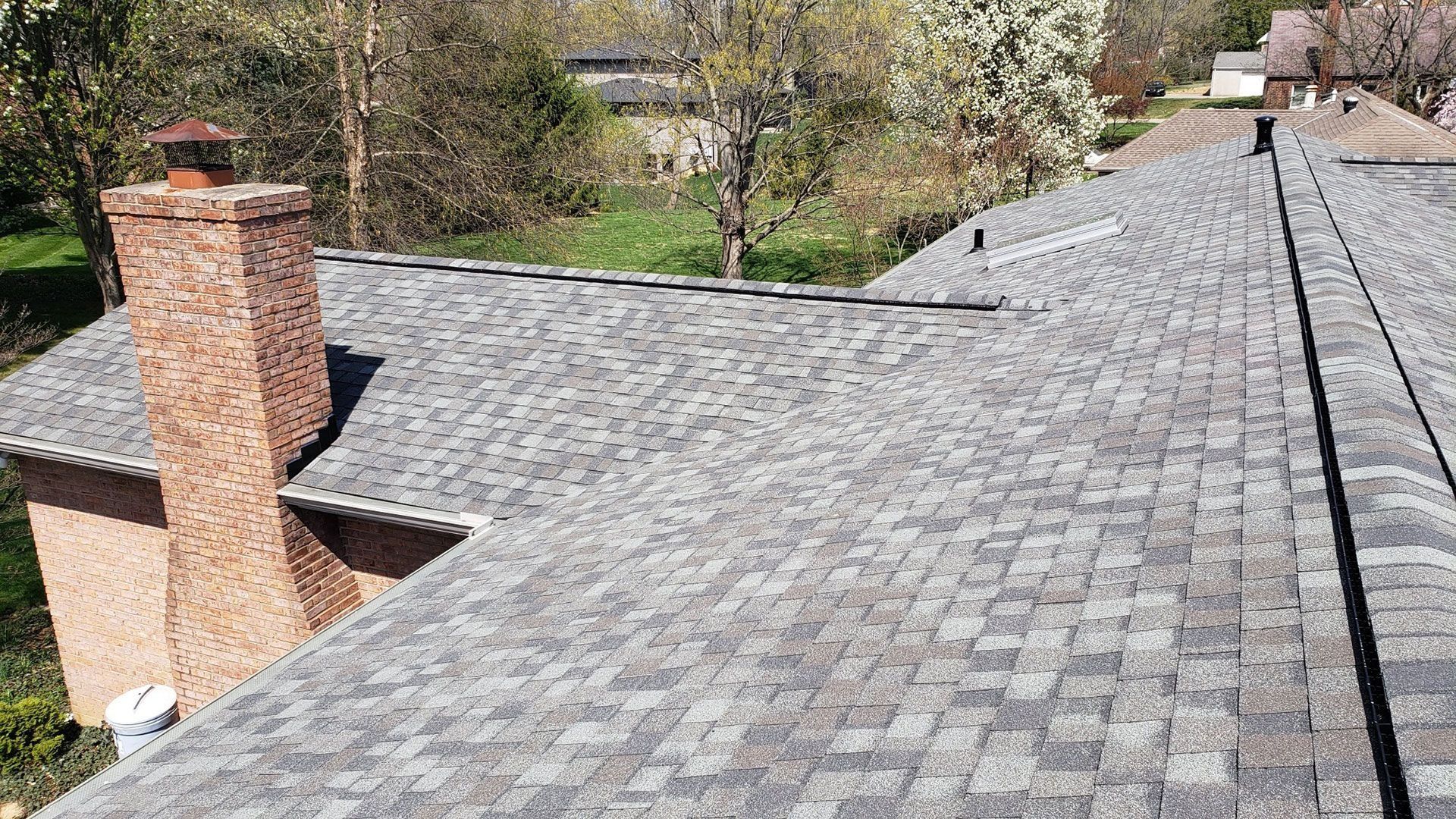 Gray shingled roof with a brick chimney and a partial view of trees and other houses.