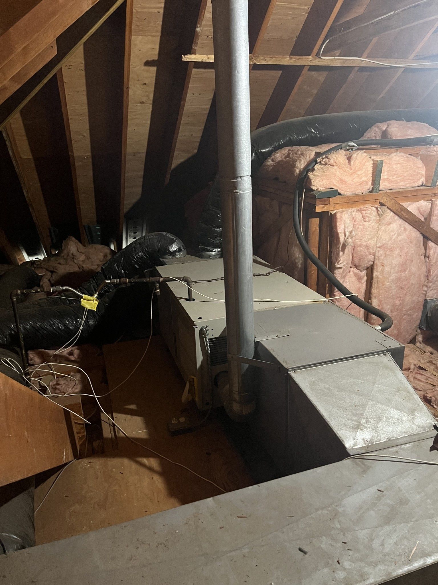 HVAC unit in an attic with insulation and ductwork. Gray metal against brown wood.