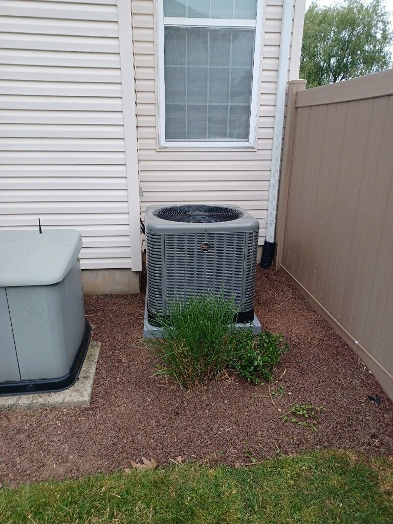 Air conditioning unit next to a fence and house with some vegetation growing at its base.