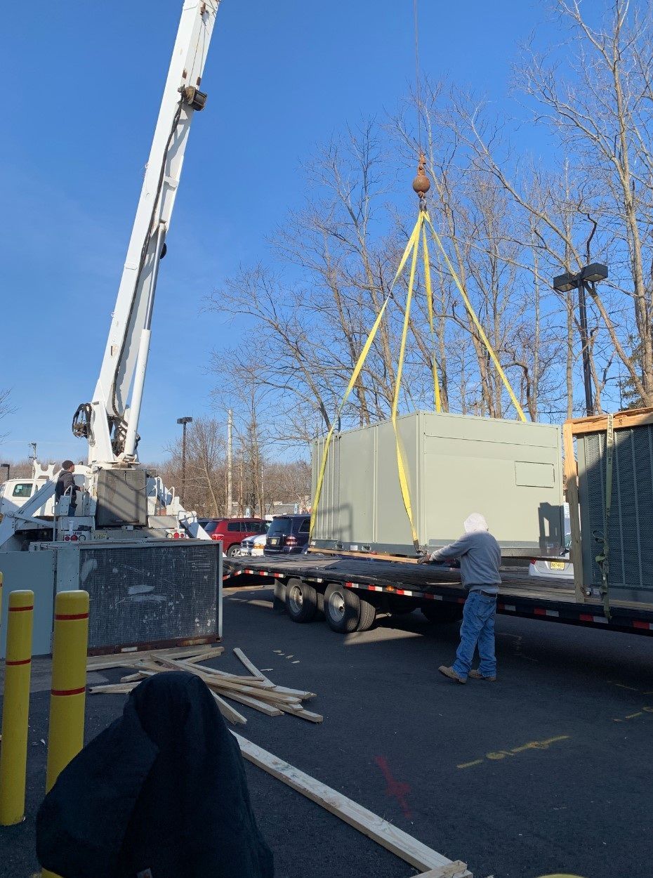 A crane lifting a large HVAC unit off a flatbed truck, worker present. Outdoors, sunny.