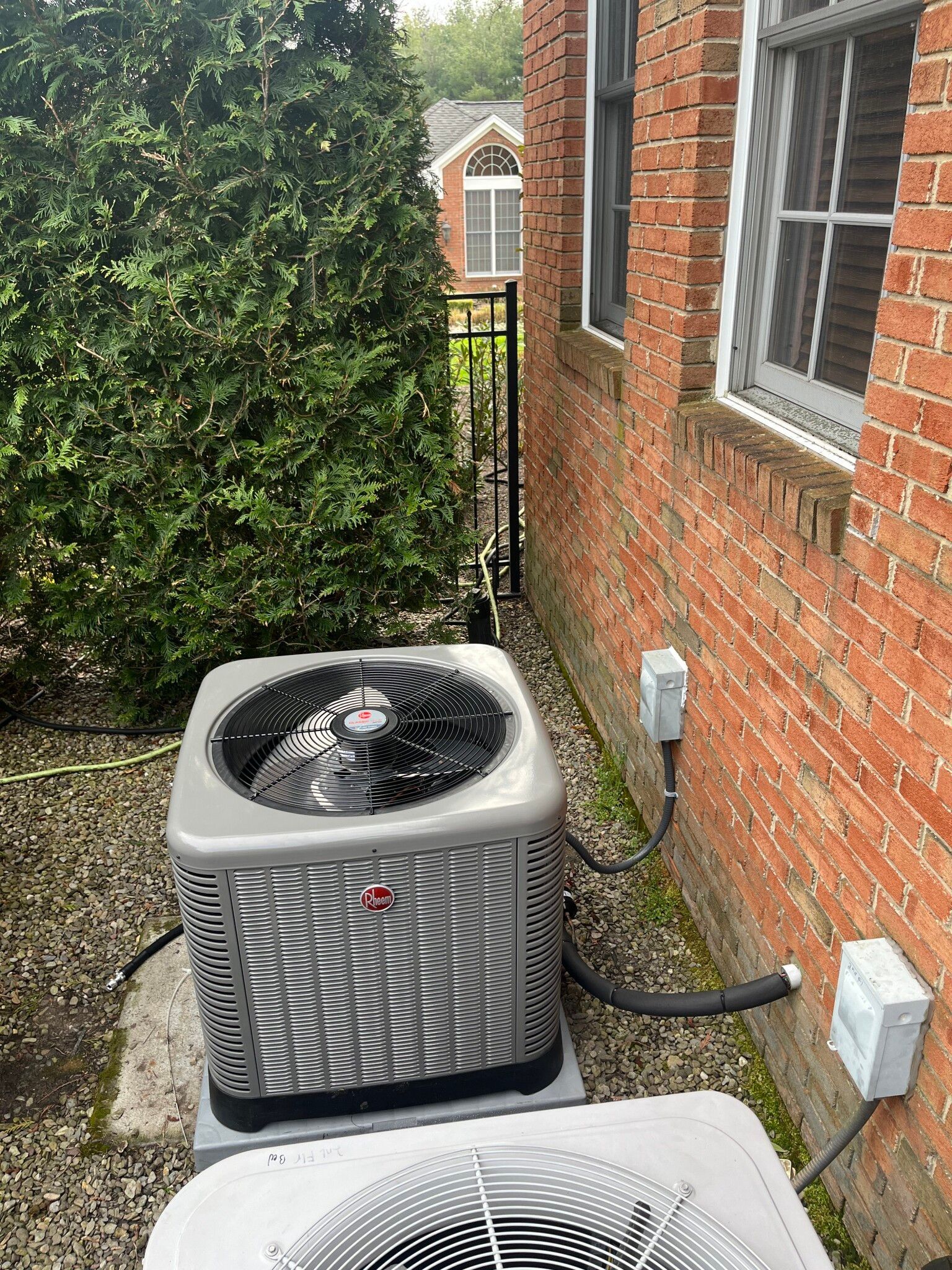 Air conditioner unit beside a brick building, next to a green bush and black fence.