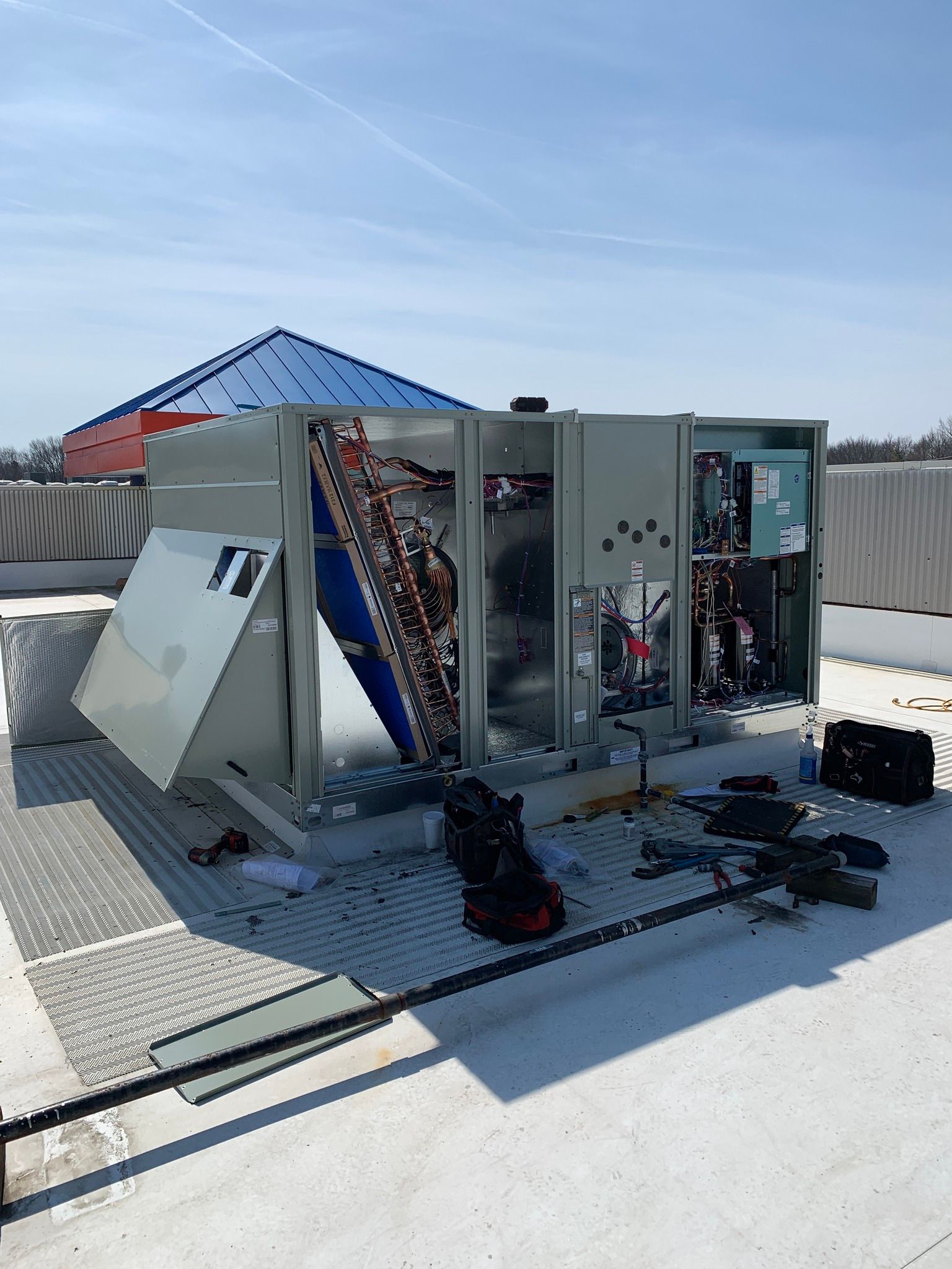 HVAC unit on a rooftop, panels open. Tools and equipment surrounding the unit on a sunny day.