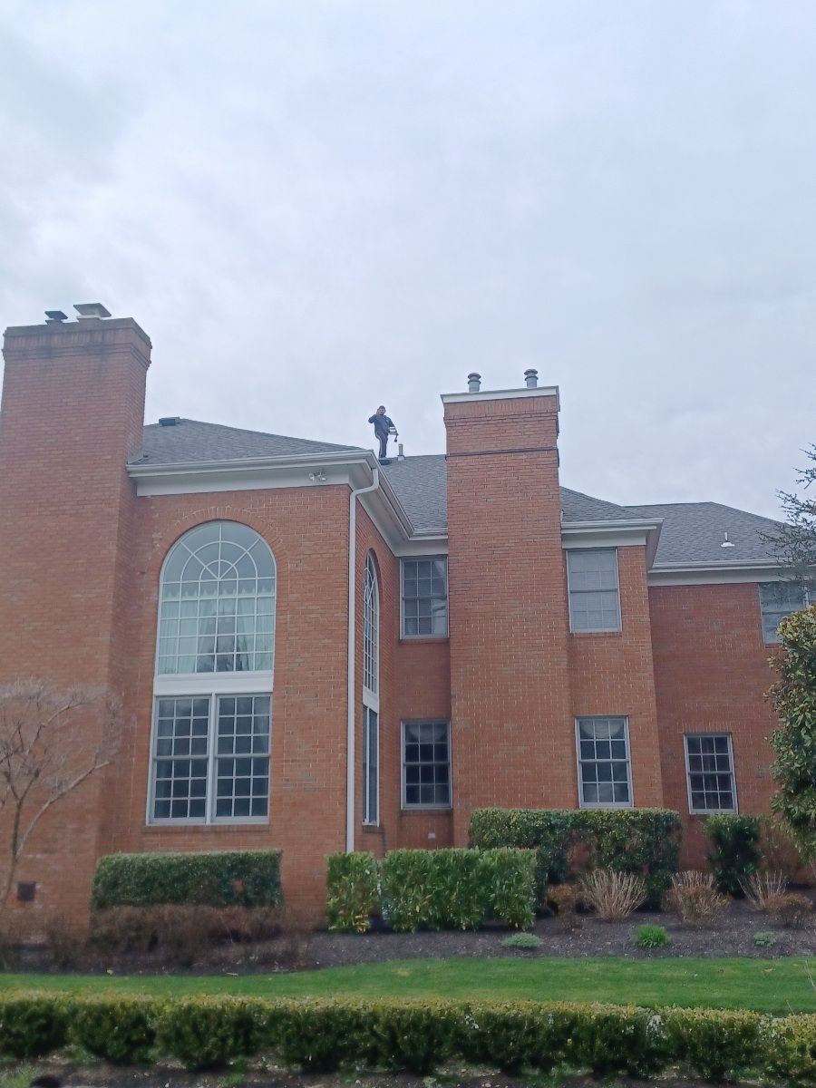 Person on a roof, cleaning the gutters of a brick two-story house with green hedges and cloudy sky.