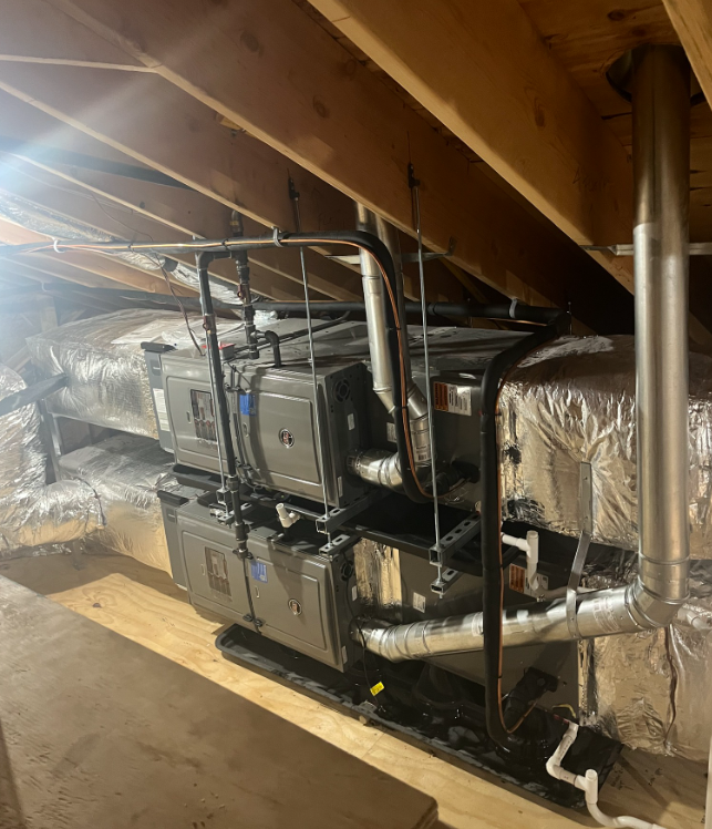 HVAC units installed in an attic space. Multiple gray boxes with ductwork and metal piping. Wooden beams are visible.
