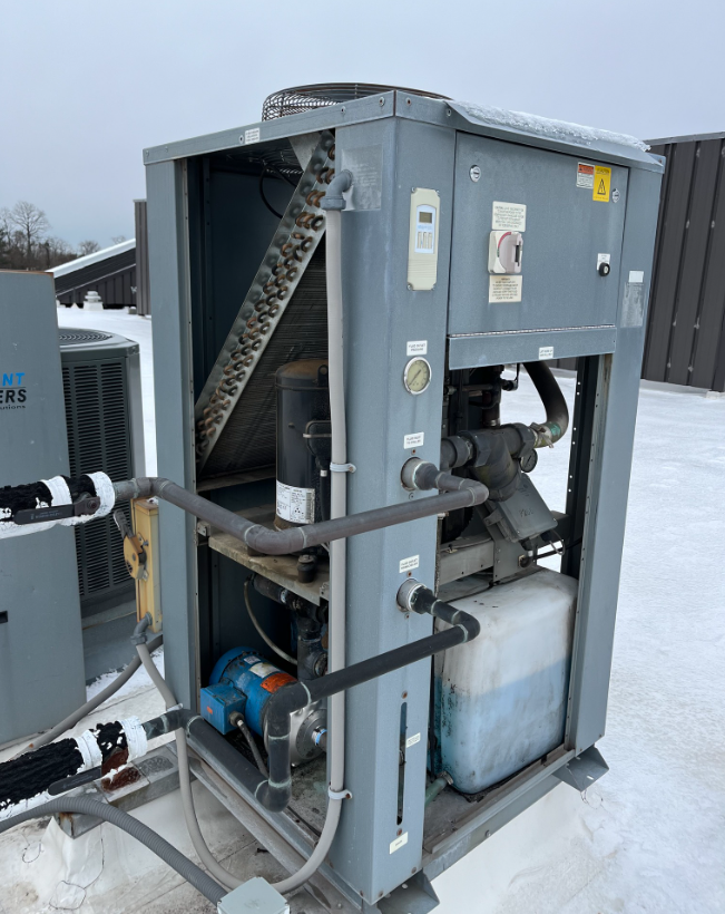 Gray industrial chiller unit on a snowy rooftop. Exposed components, pipes, and pump visible.