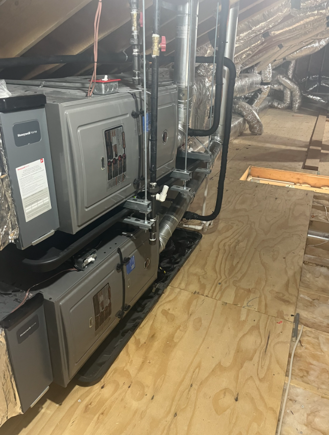HVAC system in an attic. Three grey units on wooden platform with pipes and ductwork.