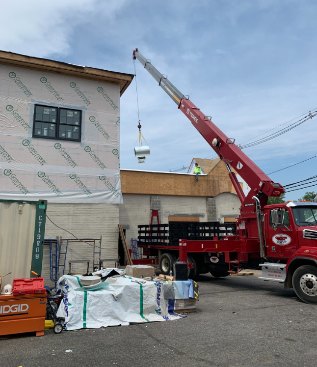 A red crane lifting an HVAC unit onto a building's roof during construction on a sunny day.