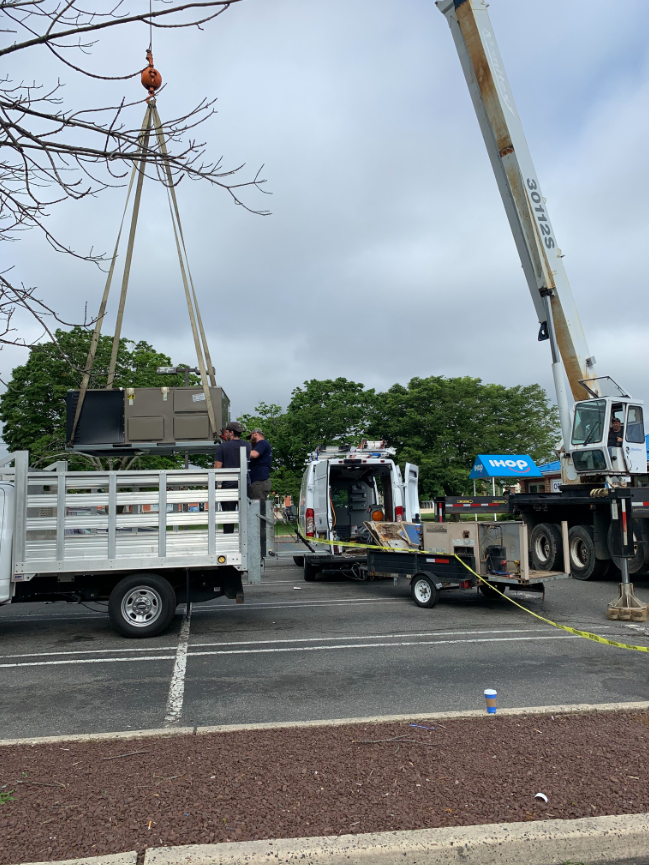 A crane lifts boxes into a truck. Man assists on the truck. Parked vehicles in a parking lot.