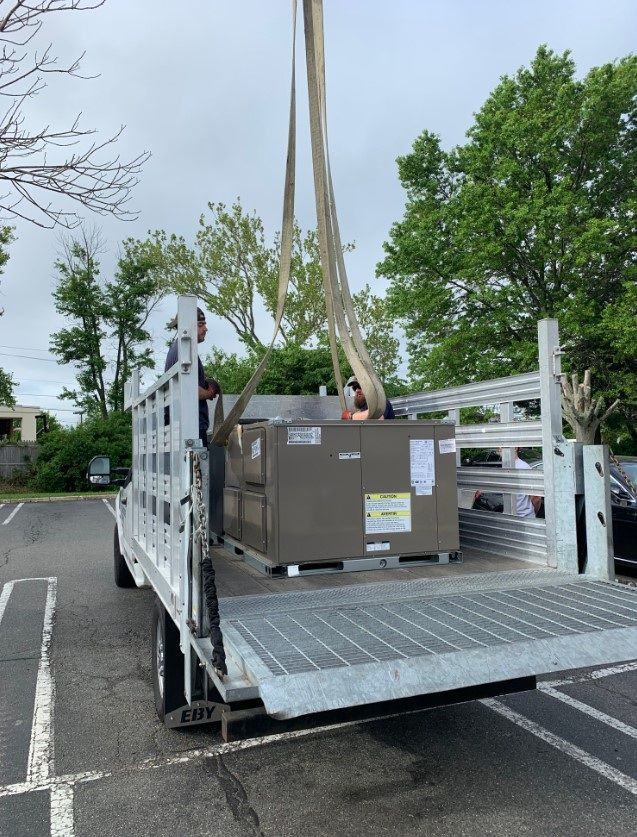 A crane lifting a HVAC unit from a flatbed truck in a parking lot.