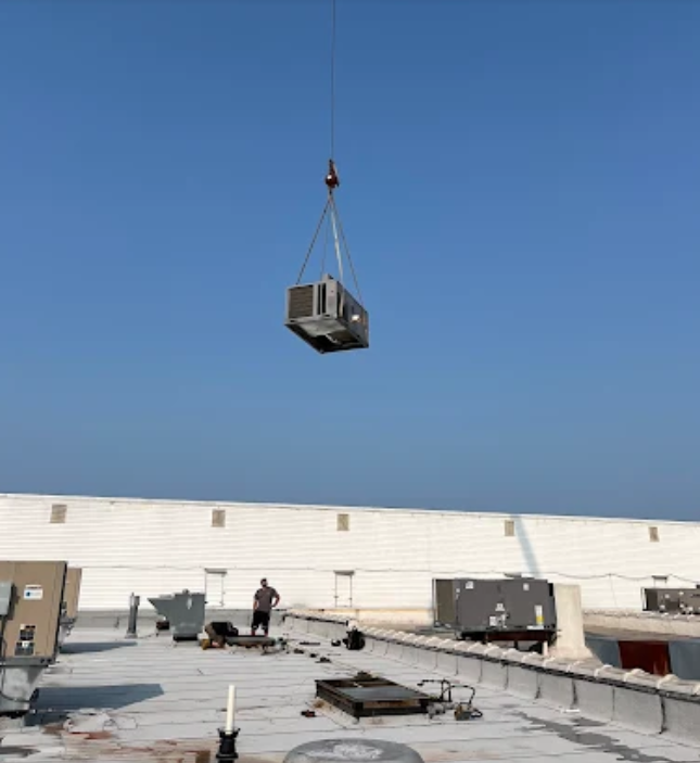 A crane lifts an air conditioning unit onto a rooftop, worker watches. Clear blue sky.