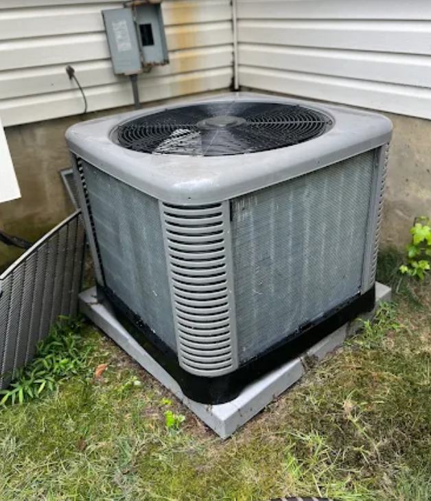 Gray air conditioner unit outside a house, on a concrete base, surrounded by grass and siding.