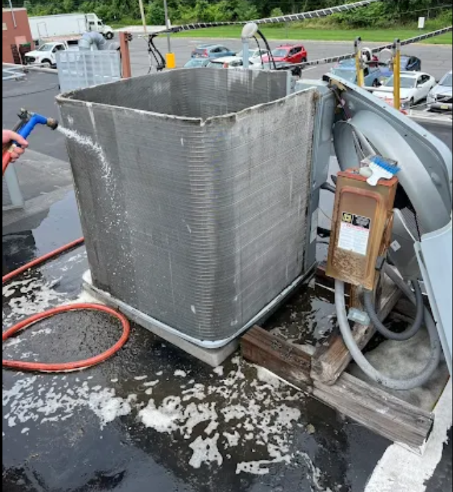 Person spraying foam cleaner on an air conditioning unit's exterior coils, outdoors on a rooftop.