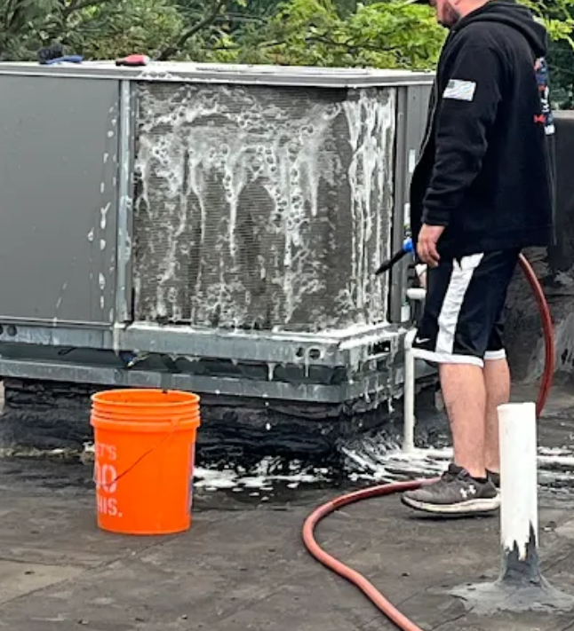 Man cleaning an AC unit with foam, orange bucket nearby. Black roof.
