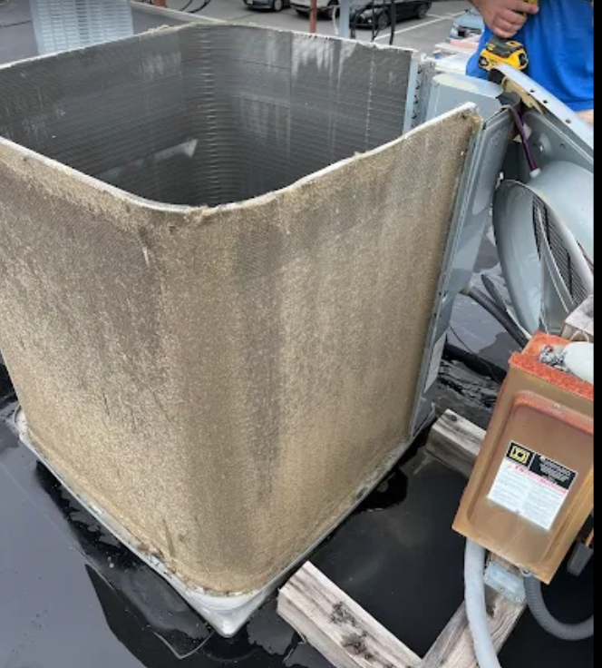 Dirty AC unit on a rooftop, being worked on. Gray metal, brown debris, and a person holding a tool.