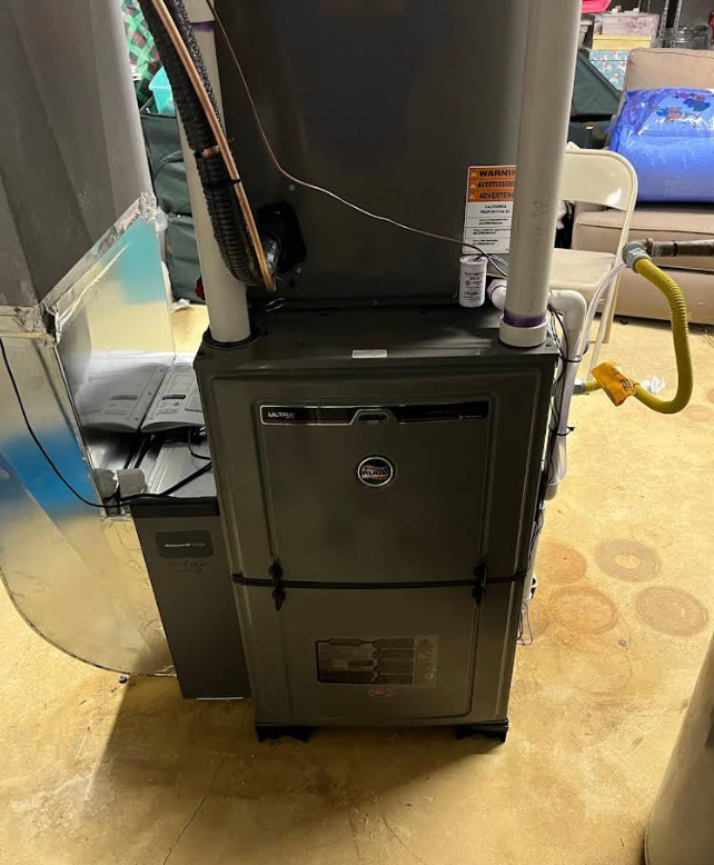 A gray furnace unit with attached pipes and vents, in a room with a yellow gas line.