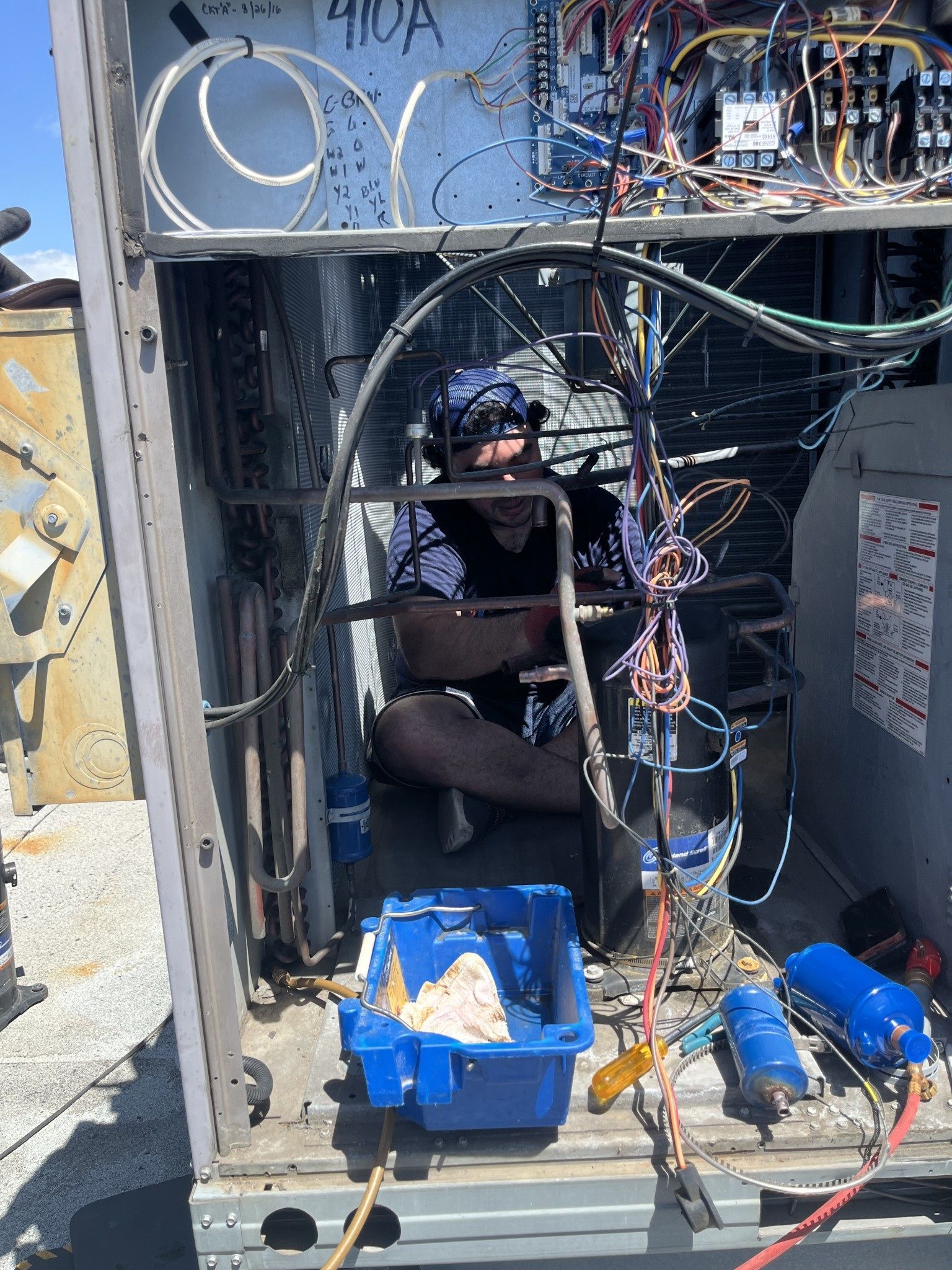 HVAC technician working inside an air conditioning unit, rooftop setting. Wires, tools, and blue toolbox visible.