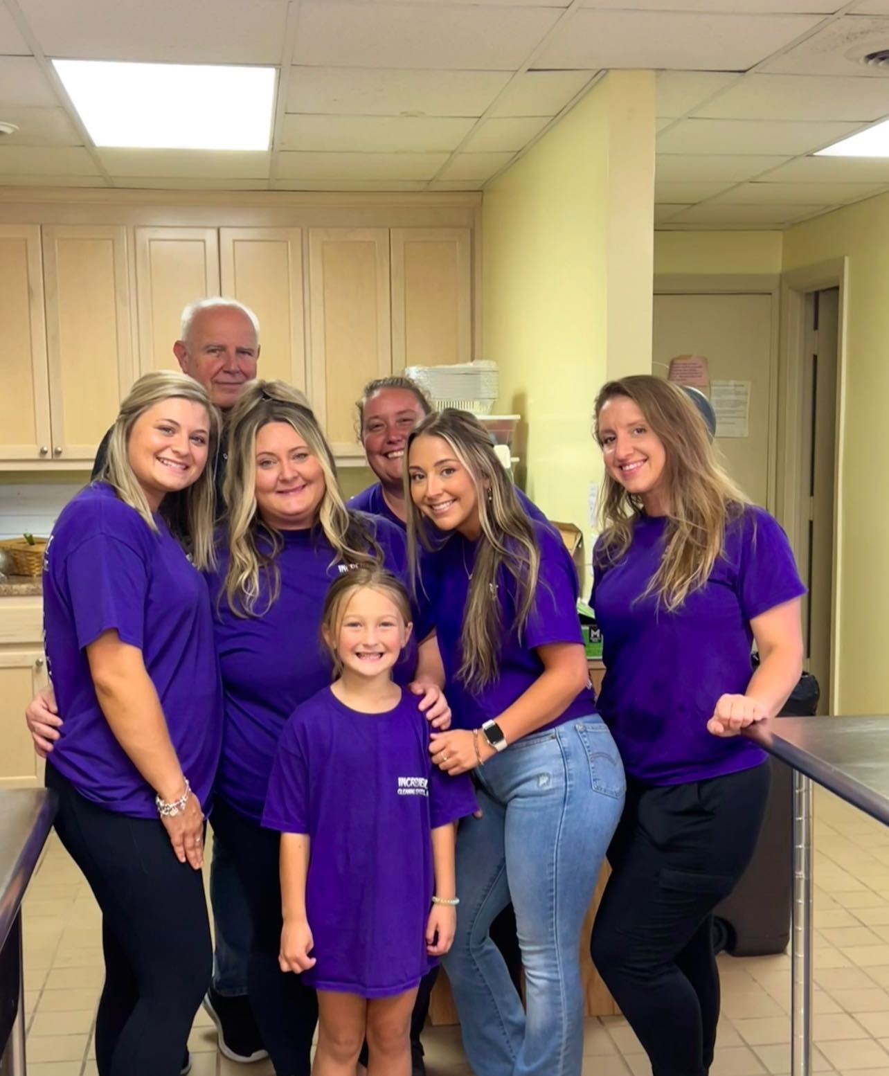 A group of people, mostly women, in purple shirts posing together in a kitchen. Smiling faces, some with arms around each other.