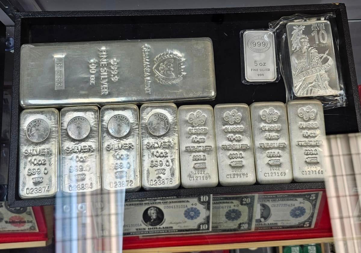 Display case of silver bars, coins, and currency notes arranged in rows.