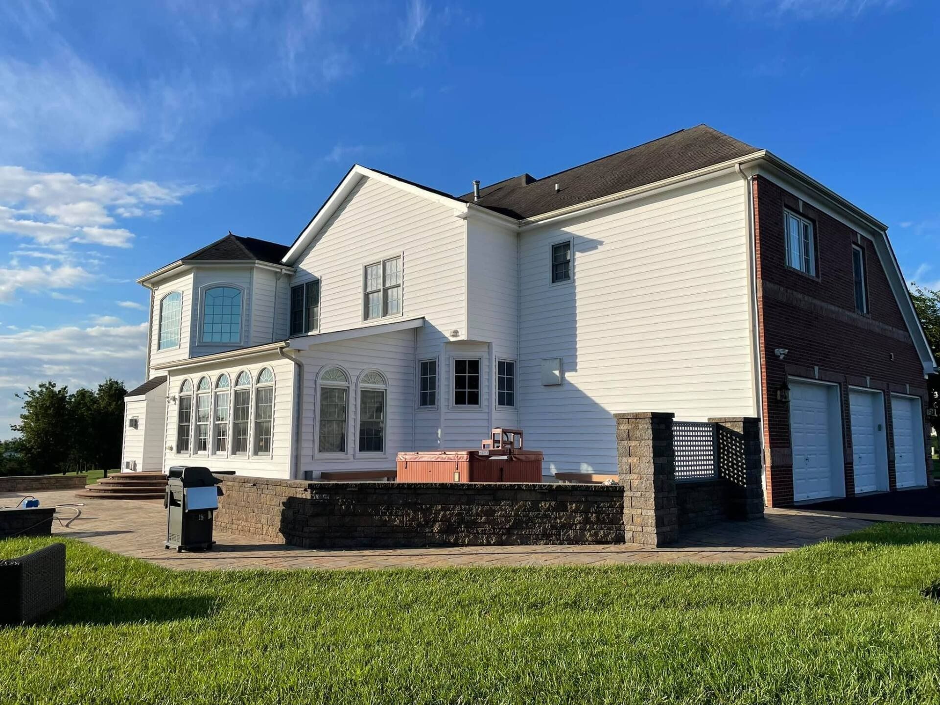 White two-story house with a brick garage, stone patio, and green grass against a blue sky.